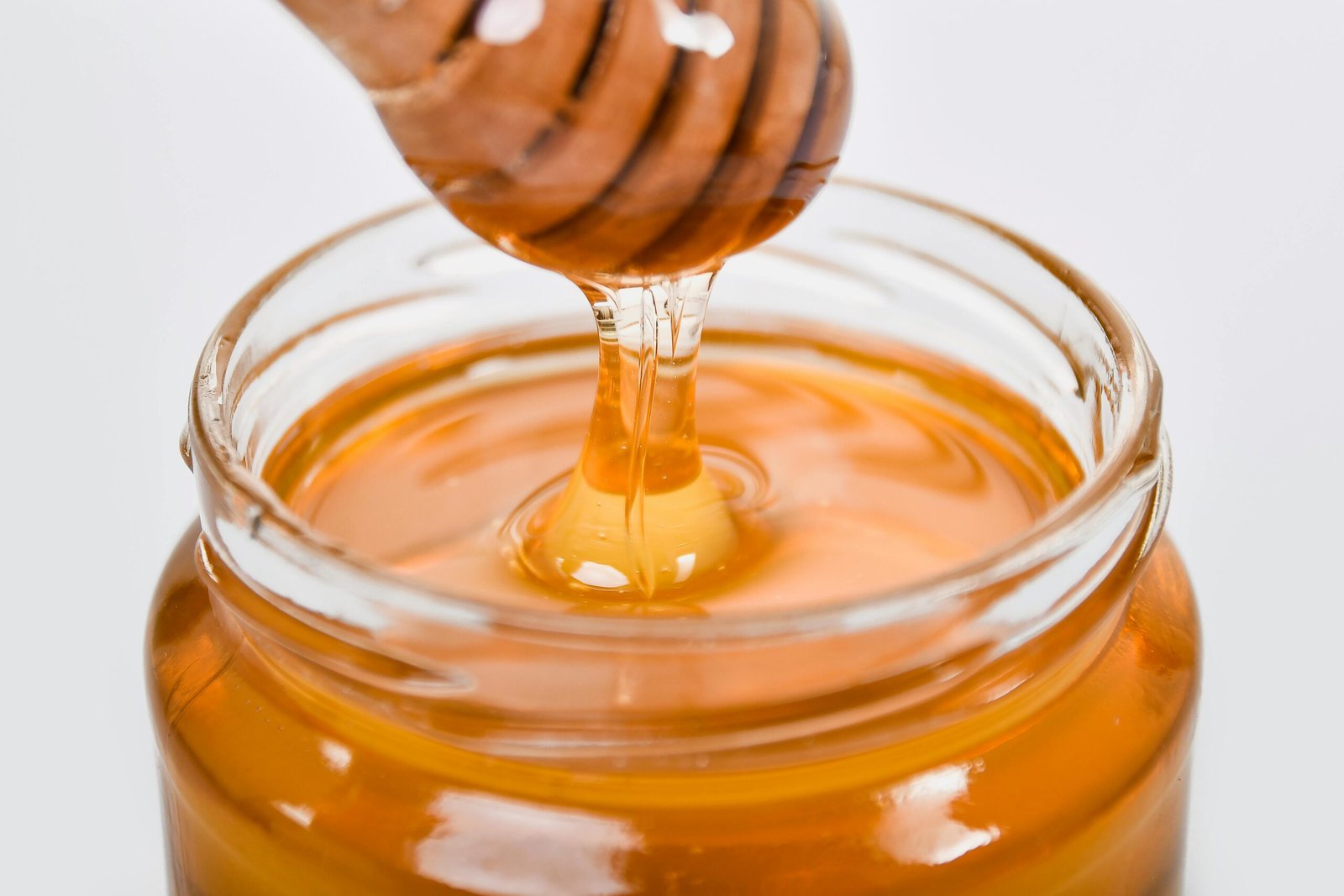 Close-up of honey flowing from a wooden dipper into a glass jar on a white background.